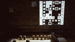photo of the installation: a row of nine bricks in the floor, all except two of them standing up. in the wall there's a projection of a monochromatic grid composed of different types of squares.